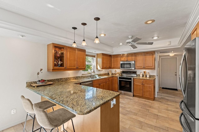 kitchen with brown cabinetry, appliances with stainless steel finishes, a peninsula, a tray ceiling, and a sink