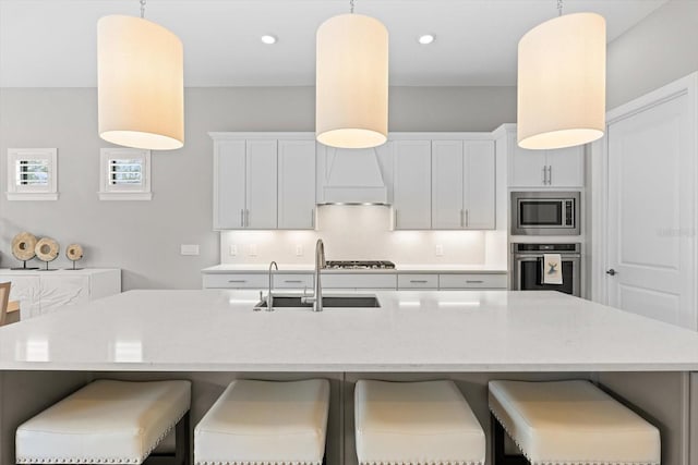 kitchen featuring premium range hood, a kitchen island with sink, a sink, appliances with stainless steel finishes, and decorative light fixtures