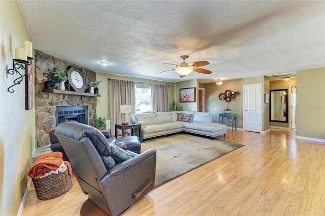 living area with a textured ceiling, wood finished floors, a stone fireplace, baseboards, and ceiling fan