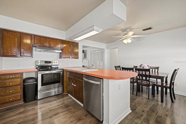 kitchen with appliances with stainless steel finishes, dark wood-style flooring, a peninsula, under cabinet range hood, and a sink