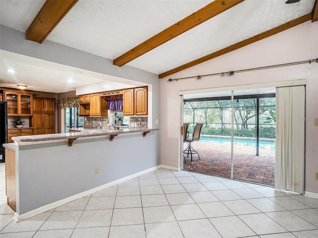 kitchen with backsplash, vaulted ceiling with beams, light tile patterned floors, brown cabinets, and freestanding refrigerator