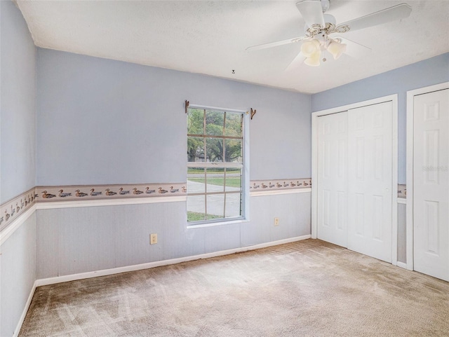 unfurnished bedroom featuring two closets, a ceiling fan, carpet floors, and wainscoting