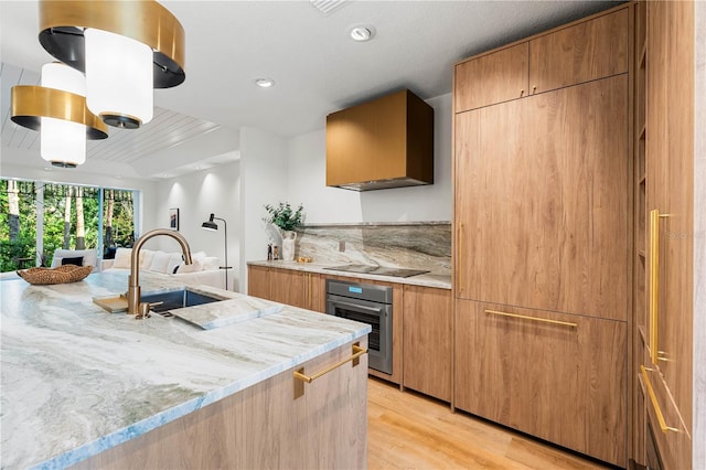 kitchen with black electric cooktop, a sink, stainless steel oven, light wood-style floors, and wall chimney exhaust hood