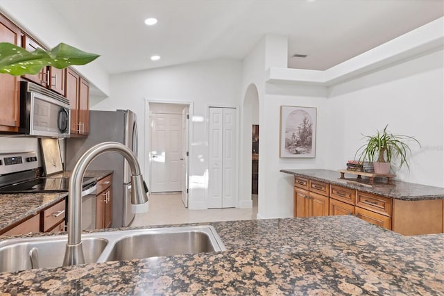 kitchen with stainless steel microwave, brown cabinetry, vaulted ceiling, a sink, and dark stone countertops