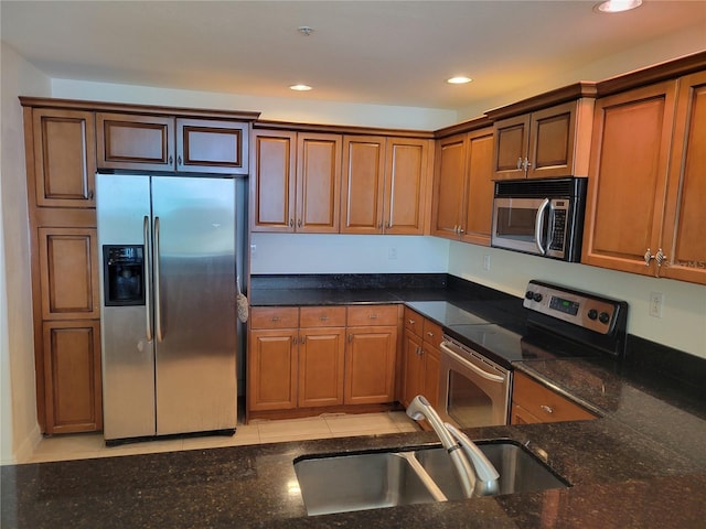 kitchen with brown cabinetry, dark stone countertops, stainless steel appliances, a sink, and recessed lighting