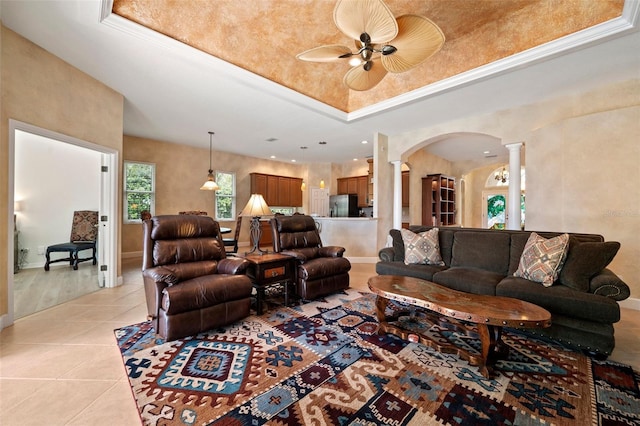 living area featuring a tray ceiling, arched walkways, light tile patterned floors, ornate columns, and baseboards