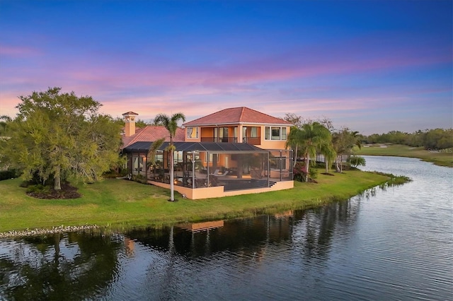 back of property at dusk featuring a water view, driveway, a yard, a lanai, and a chimney