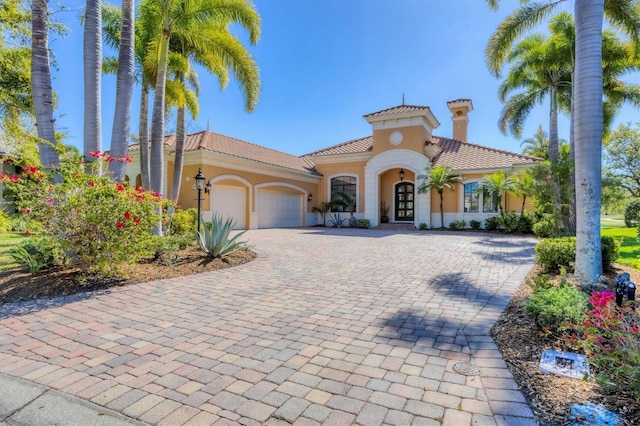 mediterranean / spanish-style house featuring a tile roof, stucco siding, a chimney, decorative driveway, and an attached garage