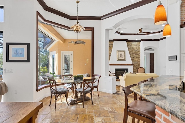 dining space featuring a large fireplace, baseboards, ceiling fan, ornamental molding, and stone tile floors