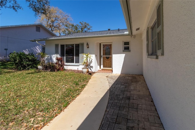 doorway to property with a shingled roof, a lawn, and stucco siding