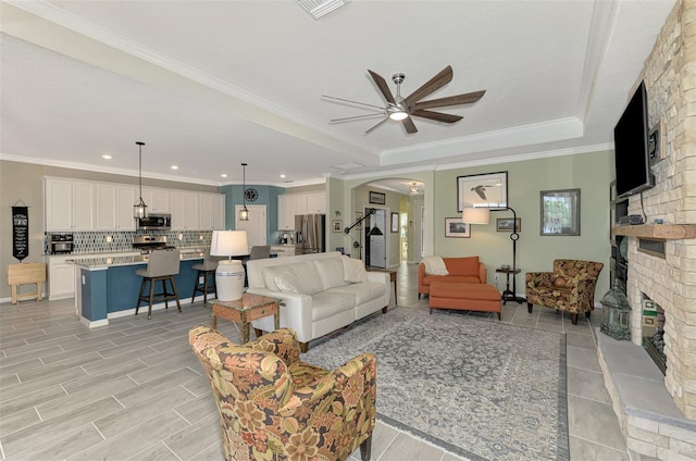 living area with arched walkways, a brick fireplace, a raised ceiling, and crown molding