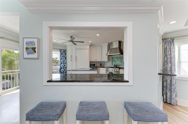 kitchen featuring crown molding, decorative backsplash, wall chimney range hood, high end stove, and a peninsula