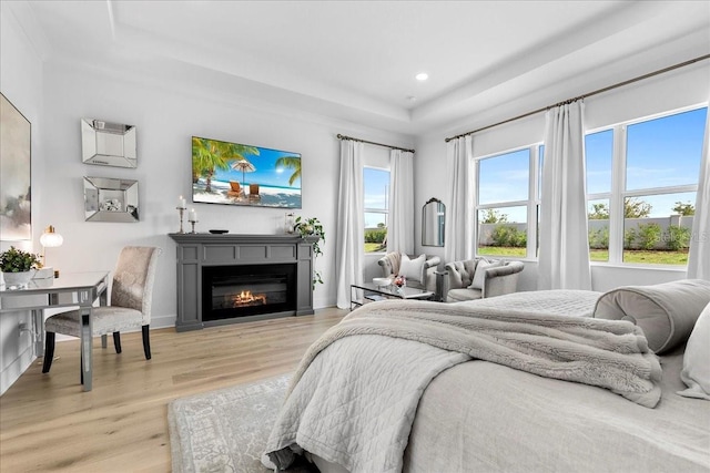 bedroom featuring light wood-type flooring, a glass covered fireplace, a tray ceiling, and multiple windows