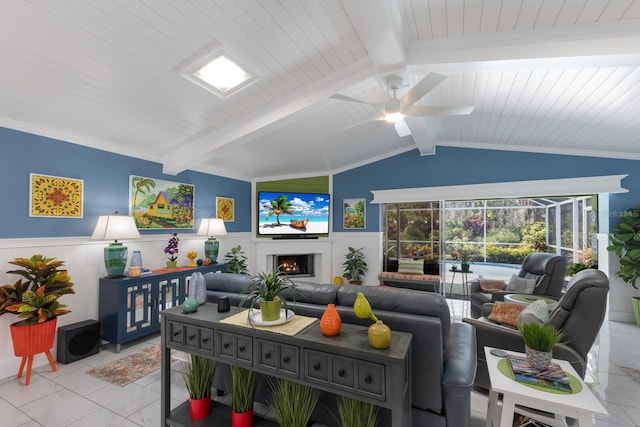 living area with a warm lit fireplace, a ceiling fan, lofted ceiling with beams, a wainscoted wall, and light tile patterned flooring