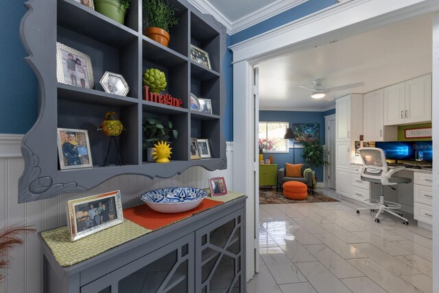 home office featuring marble finish floor, crown molding, built in desk, a ceiling fan, and wainscoting