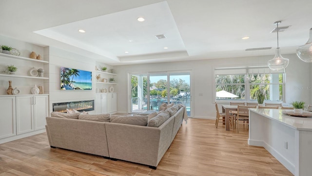 living room featuring light wood-type flooring, visible vents, built in features, a glass covered fireplace, and a raised ceiling