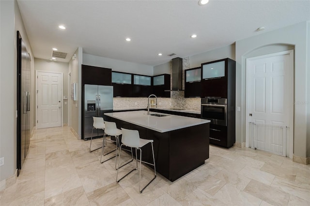 kitchen featuring wall chimney range hood, a center island with sink, light countertops, appliances with stainless steel finishes, and a sink