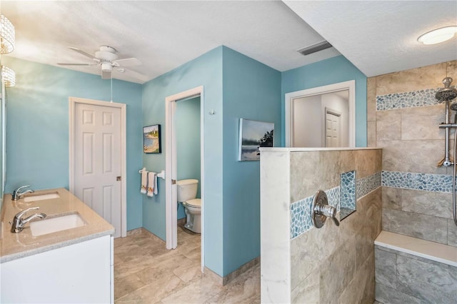 full bath with tiled shower, ceiling fan, a sink, a textured ceiling, and toilet