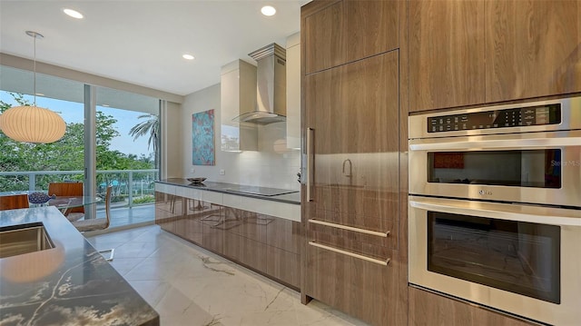 kitchen with stainless steel double oven, wall chimney range hood, brown cabinets, tasteful backsplash, and modern cabinets