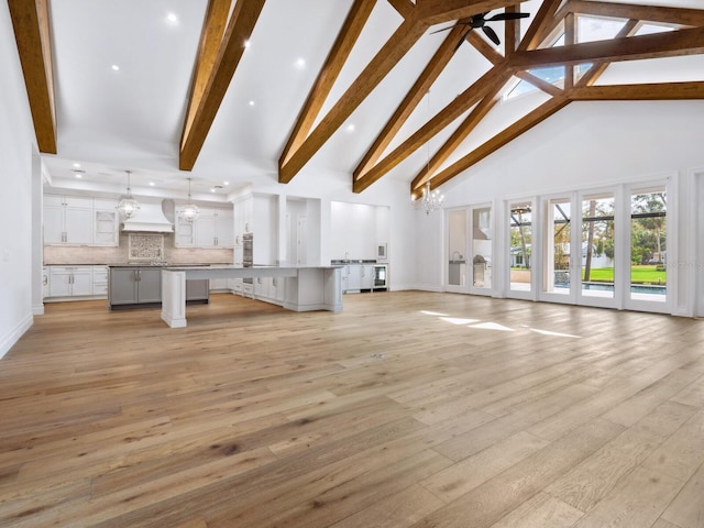 unfurnished living room featuring beam ceiling, ceiling fan with notable chandelier, high vaulted ceiling, and light wood-type flooring