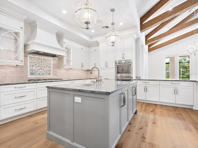 kitchen featuring vaulted ceiling with beams, an island with sink, custom range hood, appliances with stainless steel finishes, and light wood-style floors