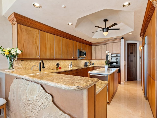 kitchen with stainless steel microwave, backsplash, a tray ceiling, a peninsula, and brown cabinetry