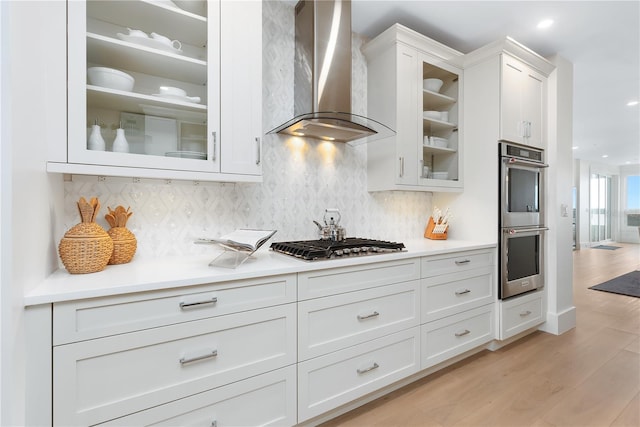 kitchen with light wood-style flooring, stainless steel appliances, white cabinetry, wall chimney range hood, and tasteful backsplash