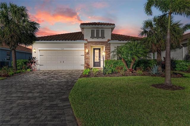 mediterranean / spanish home with stucco siding, decorative driveway, stone siding, a yard, and an attached garage