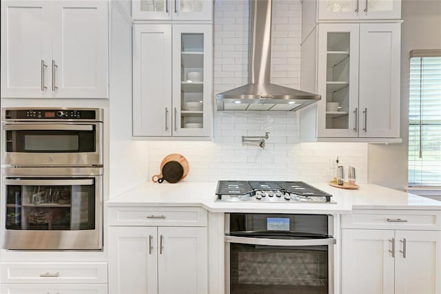 kitchen featuring decorative backsplash, wall chimney exhaust hood, glass insert cabinets, stainless steel appliances, and light countertops