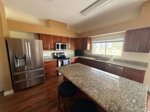 kitchen featuring stainless steel appliances, a sink, light stone countertops, dark wood-style floors, and a kitchen bar