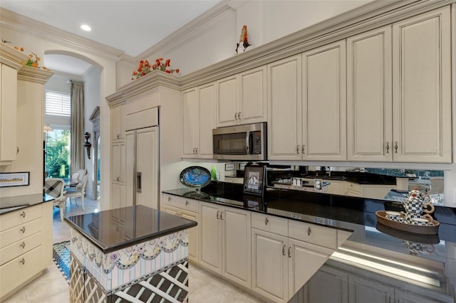 kitchen featuring ornamental molding, stainless steel microwave, dark countertops, cream cabinets, and arched walkways