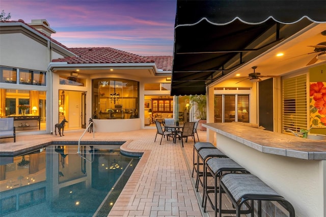 pool at dusk with a patio, a ceiling fan, an outdoor pool, and outdoor dry bar