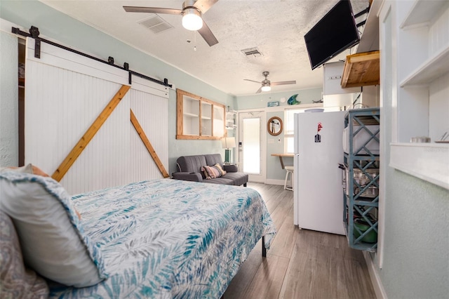 bedroom featuring wood finished floors, visible vents, freestanding refrigerator, a textured ceiling, and a barn door