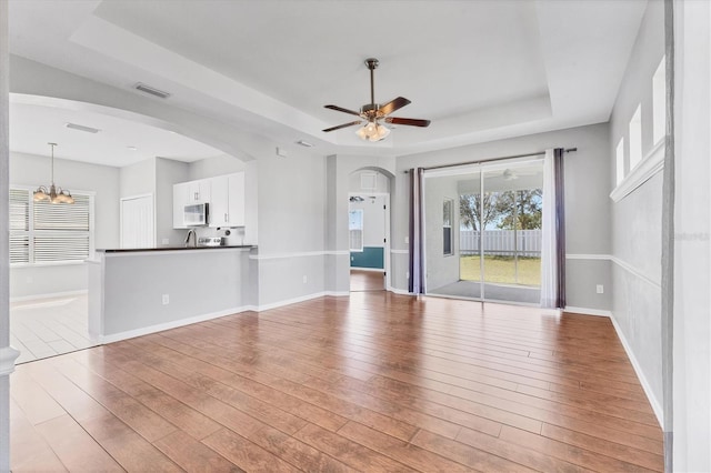 unfurnished living room featuring visible vents, ceiling fan with notable chandelier, a tray ceiling, and wood finished floors