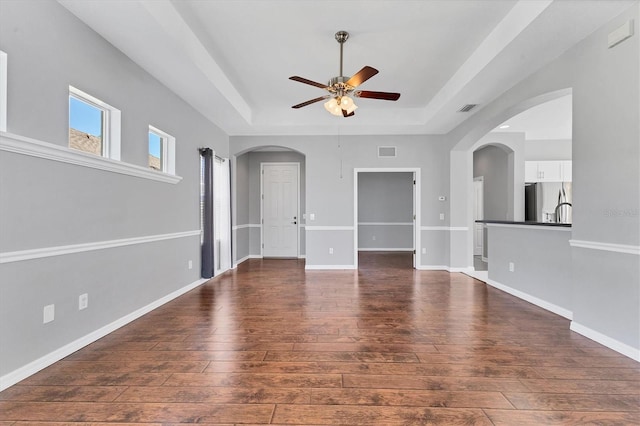 unfurnished living room with a raised ceiling, hardwood / wood-style floors, arched walkways, baseboards, and ceiling fan