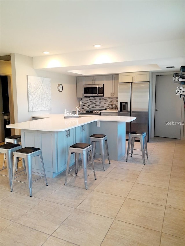 kitchen featuring gray cabinetry, light countertops, appliances with stainless steel finishes, a kitchen bar, and backsplash