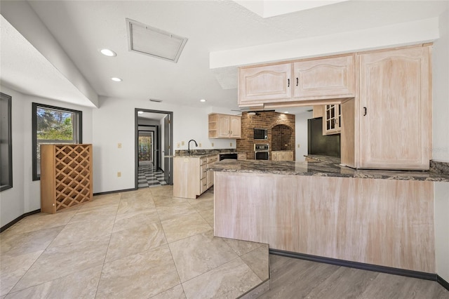 kitchen with oven, light brown cabinetry, dark stone counters, a peninsula, and a sink