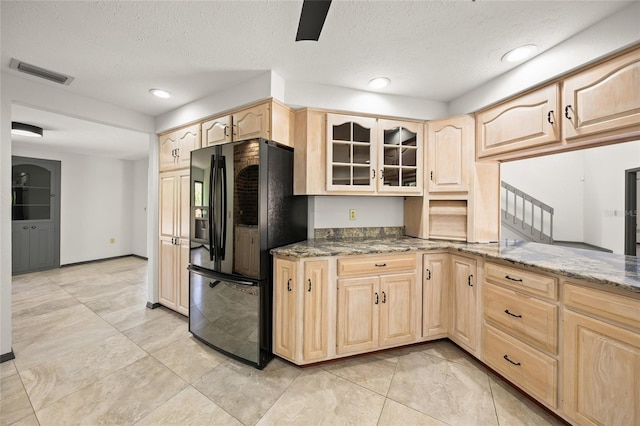 kitchen featuring visible vents, light brown cabinets, stone countertops, glass insert cabinets, and black refrigerator with ice dispenser