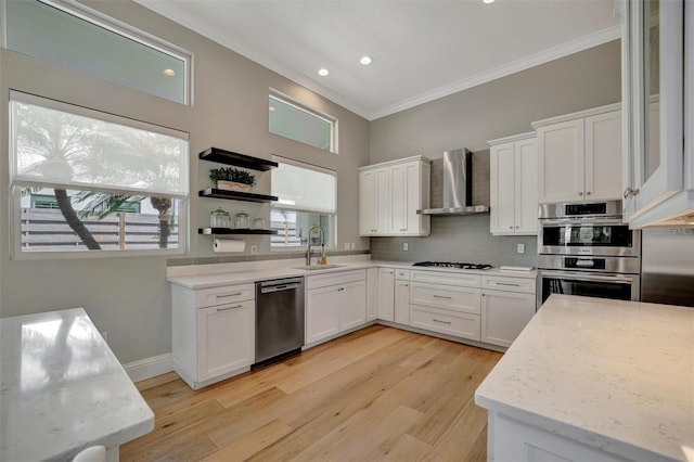 kitchen featuring crown molding, appliances with stainless steel finishes, white cabinetry, a sink, and wall chimney range hood