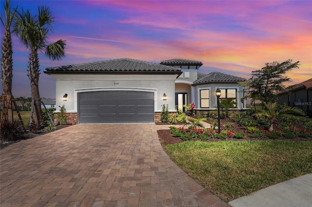 mediterranean / spanish house with stucco siding, decorative driveway, stone siding, a garage, and a tiled roof