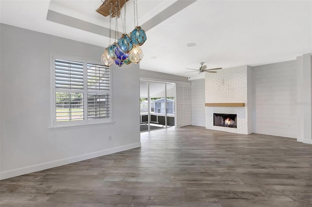 unfurnished living room featuring a ceiling fan, a tray ceiling, wood finished floors, a fireplace, and baseboards