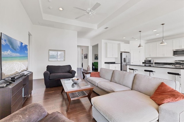 living area featuring a ceiling fan, dark wood-style floors, baseboards, a tray ceiling, and recessed lighting