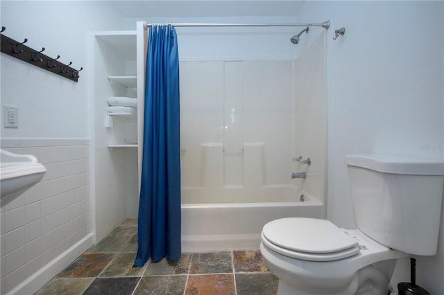 bathroom featuring stone tile flooring, toilet, shower / bath combination with curtain, and tile walls