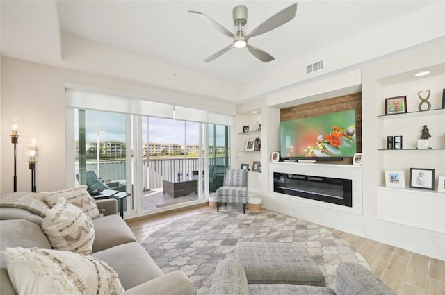 living room with visible vents, built in shelves, a glass covered fireplace, wood finished floors, and ceiling fan