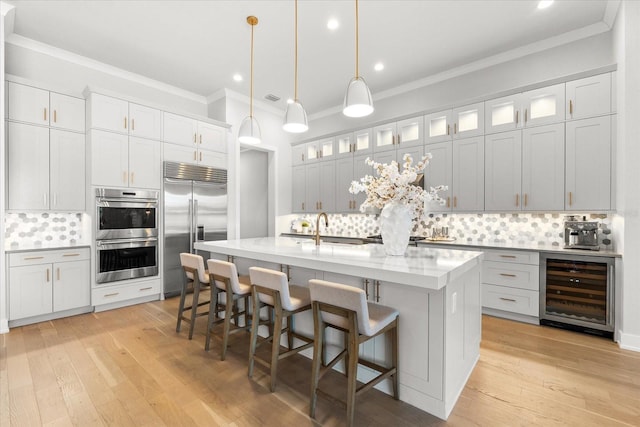 kitchen featuring light wood finished floors, a kitchen island with sink, a sink, wine cooler, and appliances with stainless steel finishes