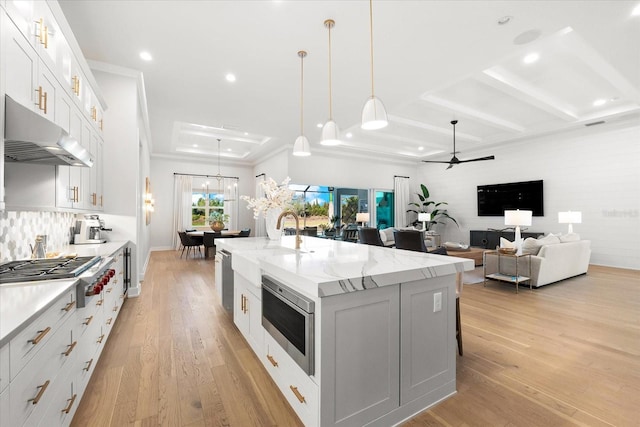 kitchen featuring oven, an island with sink, under cabinet range hood, stainless steel gas stovetop, and light wood finished floors