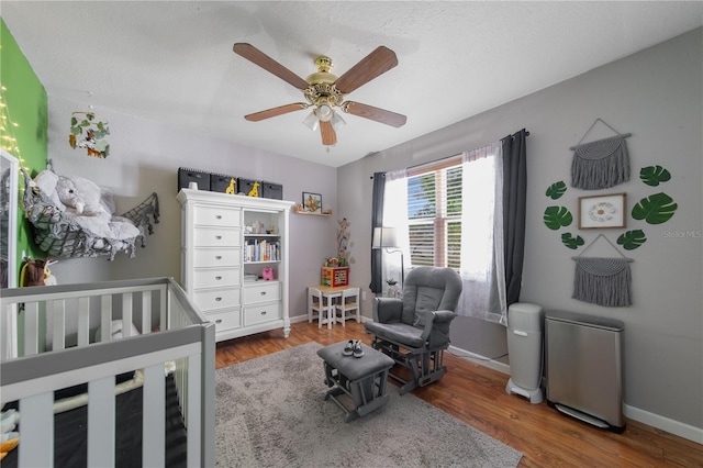 bedroom with baseboards, a nursery area, wood finished floors, and a ceiling fan