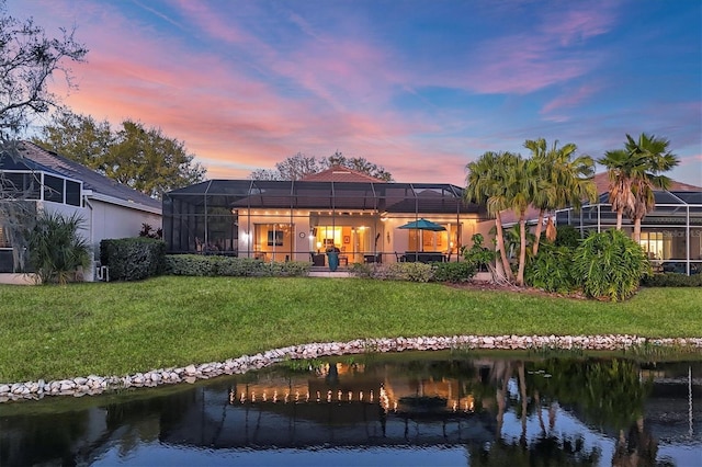 back of house at dusk featuring a lawn, glass enclosure, and a water view