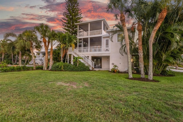 back of house at dusk featuring stucco siding, a lawn, stairs, and a sunroom