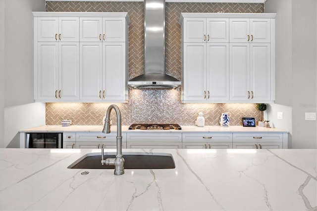 kitchen with stainless steel gas cooktop, a sink, wine cooler, white cabinets, and wall chimney exhaust hood